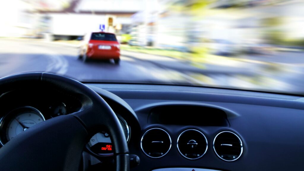 A view from the driver. Steering wheel and front console viewing a compact car on the road in front.