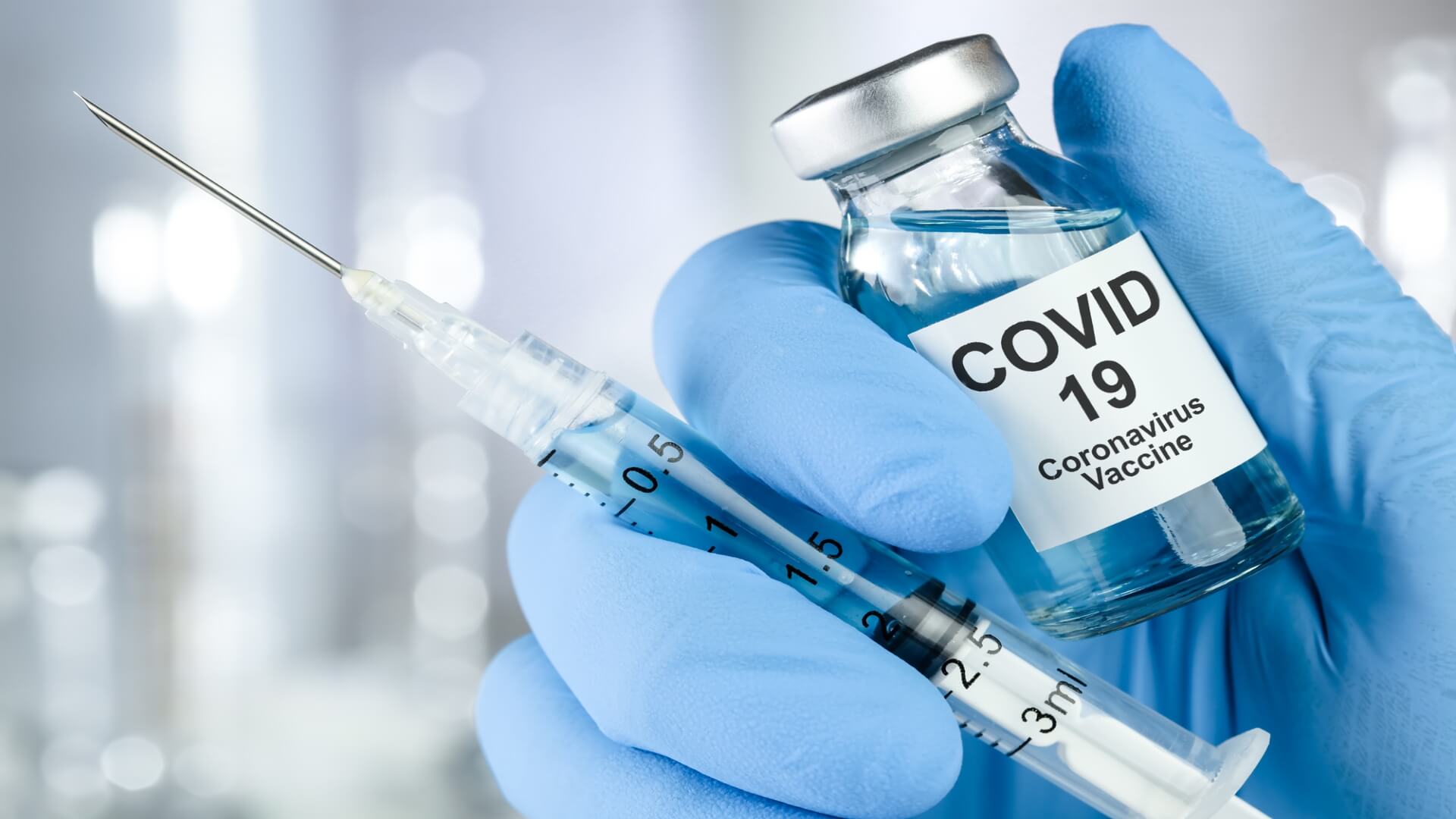 A blue gloved hand holding a syringe and small bottle of Covid 19 vaccine